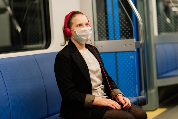 Nettes hübsches besorgtes junges Mädchen mit Maske und Kopfhörern in einem Bus, Zug oder in der U-Bahn, die zur Schule gehen?