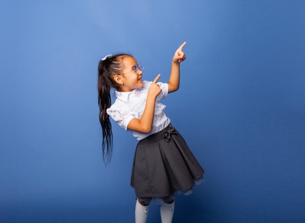 Nettes hübsches fröhliches Schulmädchen, das mit dem Finger auf den seitlichen Kopierraum zeigt, der auf blauem Hintergrund isoliert ist