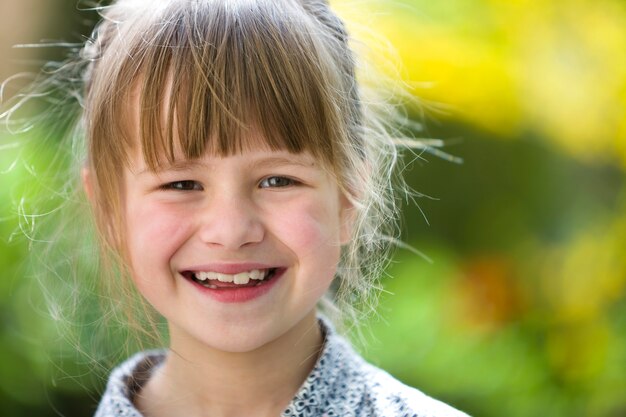 Nettes hübsches Kindermädchen mit grauen Augen und dem angemessenen Haar draußen lächelnd in camera auf unscharfer sonniger grüner und gelber heller bokeh Natur