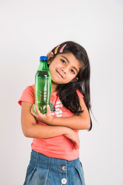 Nettes indisches oder asiatisches kleines Mädchen mit einer Haustierflasche mit Orangen- oder Mango-Kaltgetränk oder Fruchtsaft, trinken oder halten im Stehen einzeln auf blauem oder weißem Hintergrund.