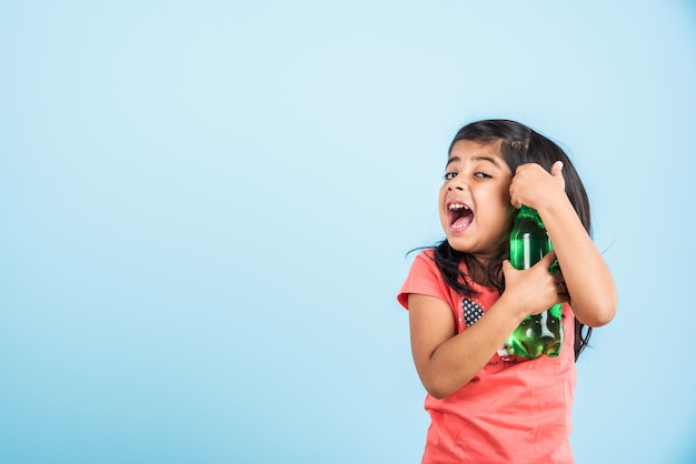 Nettes indisches oder asiatisches kleines Mädchen mit einer Haustierflasche mit Orangen- oder Mango-Kaltgetränk oder Fruchtsaft, trinken oder halten im Stehen einzeln auf blauem oder weißem Hintergrund.