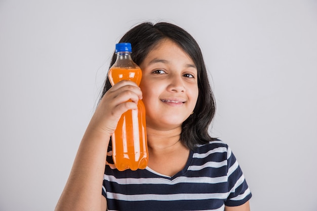 Nettes indisches oder asiatisches kleines Mädchen mit einer Haustierflasche mit Orangen- oder Mango-Kaltgetränk oder Fruchtsaft, trinken oder halten im Stehen einzeln auf blauem oder weißem Hintergrund.