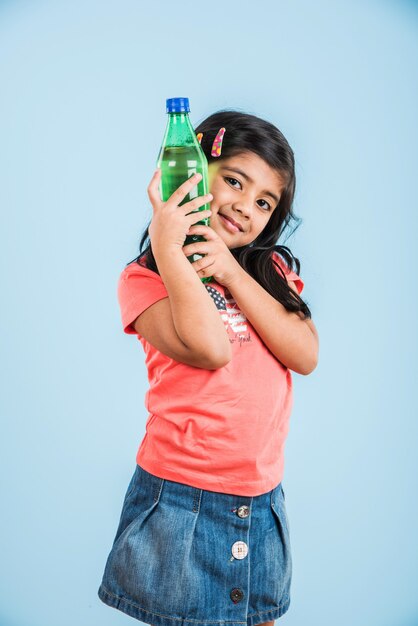 Nettes indisches oder asiatisches kleines Mädchen mit einer Haustierflasche mit Orangen- oder Mango-Kaltgetränk oder Fruchtsaft, trinken oder halten im Stehen einzeln auf blauem oder weißem Hintergrund.