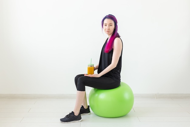 Nettes junges gemischtes Rennen-Hipster-Mädchen mit farbigem Haar, das auf einem grünen Fitball sitzt und einen Bananenprotein-Smoothie in ihren Händen auf einem weißen Hintergrund hält. Konzept für gesunde Ernährung und Bewegung. Exemplar