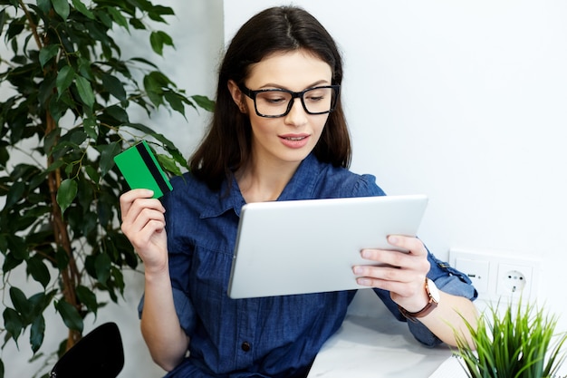 Nettes junges Mädchen, das blaues Hemd und Brillen sitzt, die mit Tablette, freiberuflichem Konzept, Online-Shopping, Halten einer Kreditkarte, Porträt sitzen.