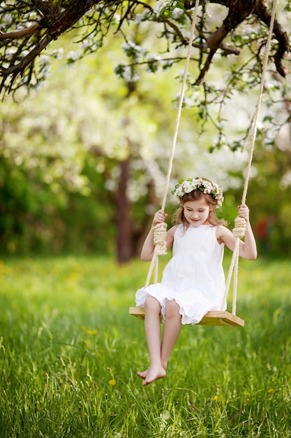 Nettes junges Mädchen, das Spaß auf einer Schaukel im blühenden alten Apfelbaumgarten hat. Sonniger Tag. Frühlingsaktivitäten im Freien für Kinder