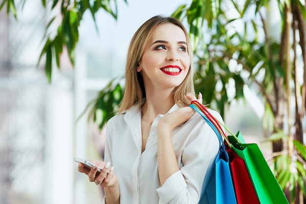 Nettes junges Mädchen mit hellbraunem Haar und roten Lippen, die weiße Bluse tragen und mit bunten Einkaufstaschen stehen, die Handy, Einkaufskonzept halten.