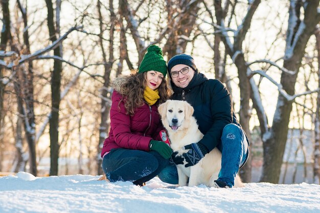 Nettes junges Paar, das Spaß im Winterpark mit ihrem Hund Golden Retriever an einem sonnigen Tag und Lächeln hat