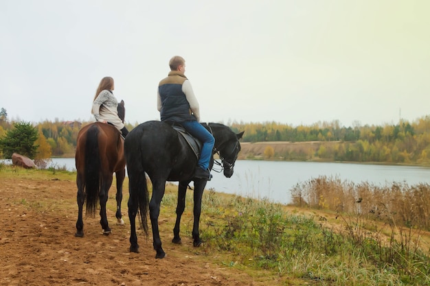 Nettes junges Paar zu Pferd im Herbstwald am See. Reiter im Herbstpark bei schlechtem bewölktem Wetter mit leichtem Regen. Konzept des Reitens, des Sports und der Erholung im Freien. Platz kopieren