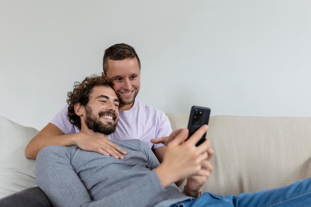 Nettes junges schwules Paar Video, das ihre Freunde in ihrem Wohnzimmer zu Hause anruft Zwei männliche Liebhaber lächeln fröhlich, während sie ihre Freunde auf einem Smartphone begrüßen Junges schwules Paar, das zusammen sitzt