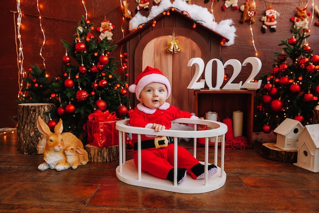 Nettes Kind, das als Weihnachtsmann verkleidet ist, sitzt in der Nähe des Weihnachtsbaums. Waren für Kinder und Urlaub. Winter-Neujahr-Konzept. Weihnachten 2022