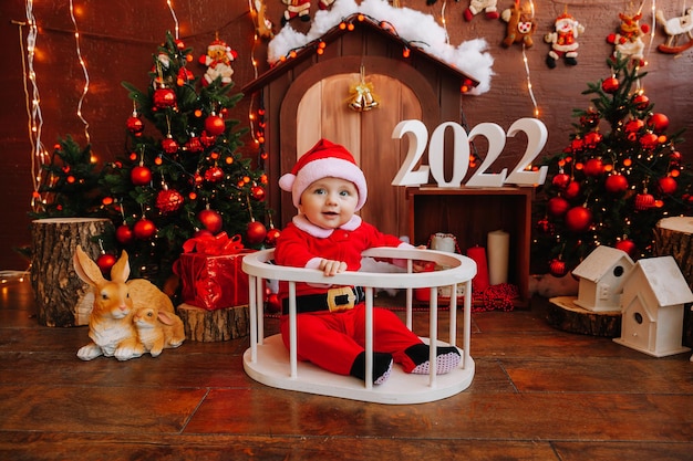 Nettes Kind, das als Weihnachtsmann verkleidet ist, sitzt in der Nähe des Weihnachtsbaums. Waren für Kinder und Urlaub. Winter-Neujahr-Konzept. Weihnachten 2022