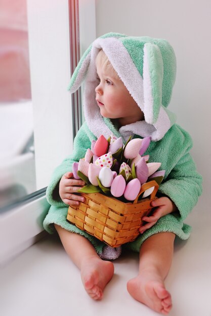 Nettes Kind in einem Osterhasen-Kostüm mit einem Blumenstrauß sitzt auf einem Fensterbrett gegen das Fenster und lächelt.