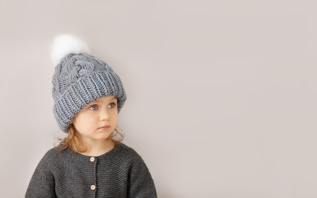 Nettes Kindermädchen in grauer handgemachter Wollstrickwintermütze mit weißem Pompon und Strickpullover posiert auf Studiohintergrund