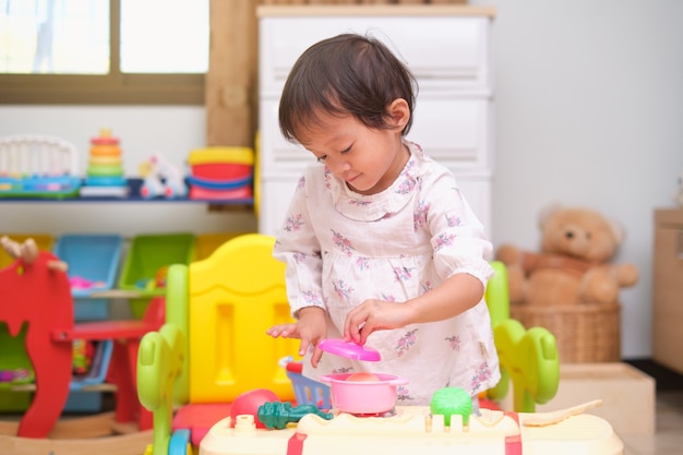 Nettes kleines asiatisches 2 - 3 Jahre altes Kleinkindmädchenkind, das Spaß hat, allein mit kochendem Spielzeug zu spielen, Küche zu Hause eingestellt