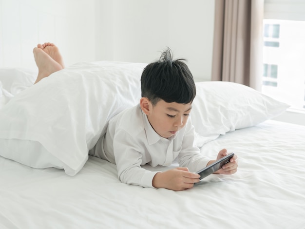 Nettes kleines asiatisches Kind konzentrierte sich auf Smartphone beim Lügen auf dem Bett