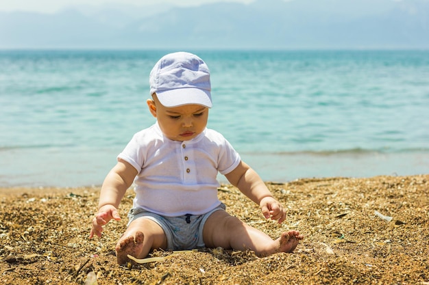 Nettes kleines Baby, das auf dem Sandstrand in der Nähe des blauen Meeres sitzt