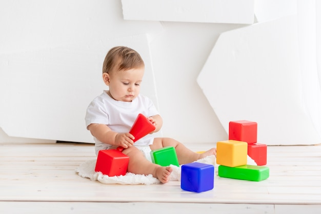 Nettes kleines Baby in einem weißen T-Shirt und Windeln, die zu Hause auf einer Matte mit bunten Würfeln spielen