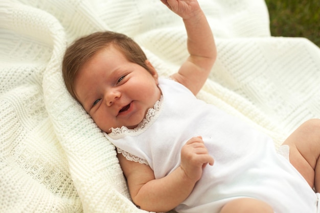 Nettes kleines Baby in weißer Kleidung, das auf der weißen Decke auf dem Gras lächelt