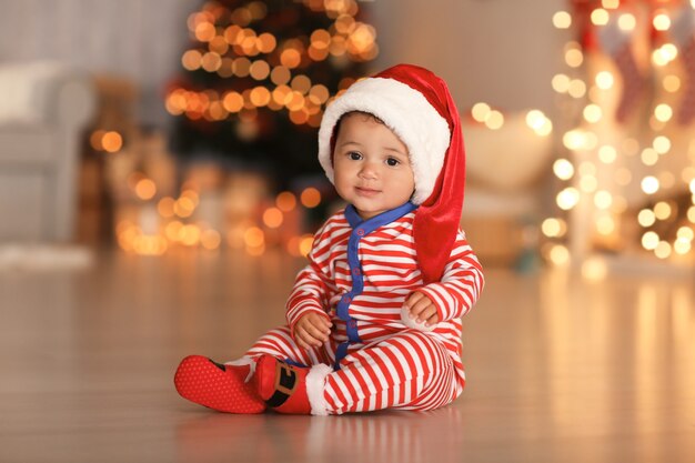 Nettes kleines Baby mit Weihnachtsmütze auf dem Boden und verschwommener Weihnachtsbeleuchtung auf der Oberfläche