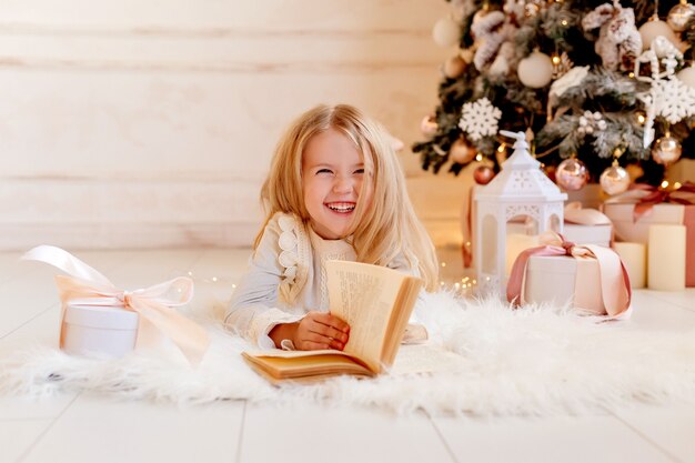 Nettes kleines blondes Mädchen liest ein Buch zu Hause nahe dem Weihnachtsbaum