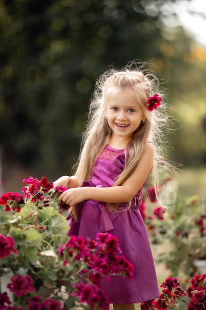 Nettes kleines blondes Mädchen mit langen Haaren mit Blumen im Sommer Pelargonium Geranium Kind lächelt Blumen pflanzen im sonnigen Garten Kleines Mädchen Gärtner Familie mit Kindern arbeiten im Hinterhof