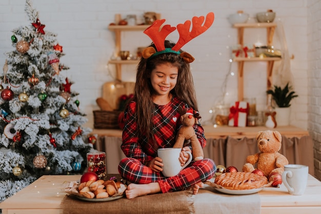 Nettes kleines brünettes Mädchen im roten Pyjama mit Rentierhörnern auf dem Kopf isst einen Weihnachtskuchen