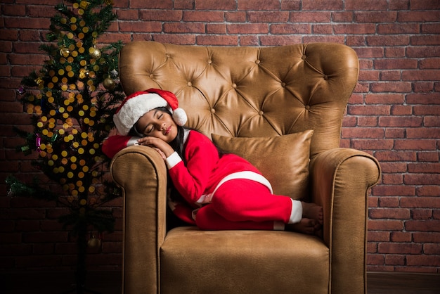 Nettes kleines indisches asiatisches Mädchen, das Weihnachten feiert, während es ein Weihnachtsmann-Kleid und einen Hut trägt, über dem Sofa mit Geschenken und einem Baum im Hintergrund gegen eine rote Backsteinmauer sitzt, verträumte Beleuchtung