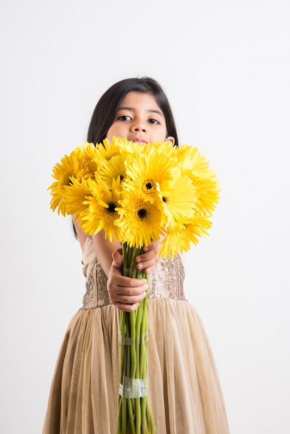 Nettes kleines indisches Mädchen, das einen Haufen oder einen Blumenstrauß frischer gelber Gerberablumen hält. Auf weißem Hintergrund isoliert