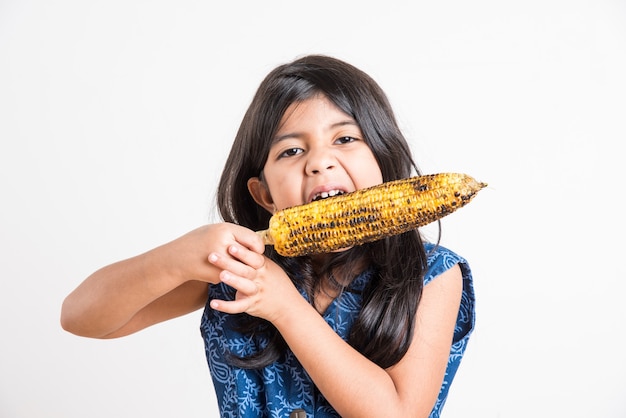 Nettes kleines indisches Mädchen, das gegrillten Mais oder Bhutta isst, stehend isoliert auf weißem Hintergrund