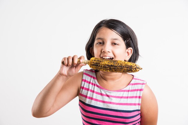 Nettes kleines indisches Mädchen, das gegrillten Mais oder Bhutta isst, stehend isoliert auf weißem Hintergrund