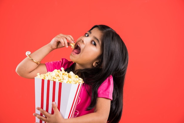 Nettes kleines indisches Mädchen, das Popcorn isst, während es eine Sonnenbrille oder eine 3D-Brille in einem Theater trägt. Stehend über buntem Hintergrund isoliert