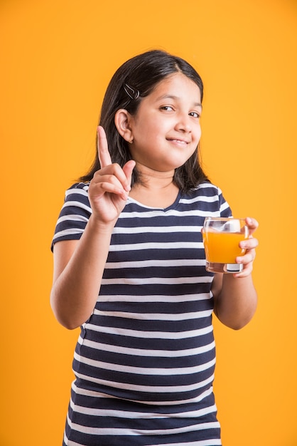 Nettes kleines indisches oder asiatisches verspieltes Mädchen, das frischen Mango- oder Orangensaft oder kaltes Getränk oder Getränk in einem Glas trinkt, einzeln auf weißem Hintergrund