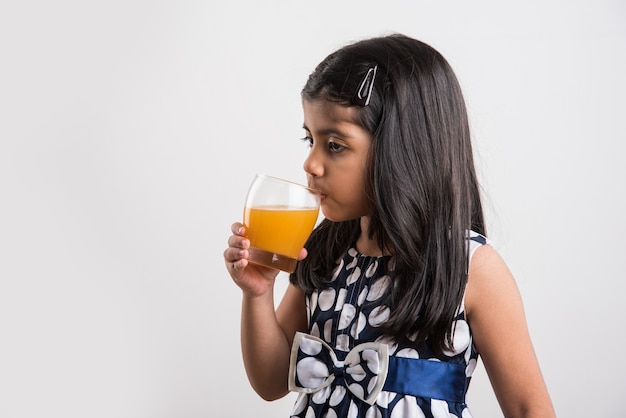 Nettes kleines indisches oder asiatisches verspieltes Mädchen, das frischen Mango- oder Orangensaft oder kaltes Getränk oder Getränk in einem Glas trinkt, einzeln auf weißem Hintergrund