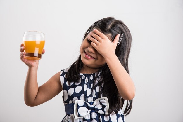 Nettes kleines indisches oder asiatisches verspieltes Mädchen, das frischen Mango- oder Orangensaft oder kaltes Getränk oder Getränk in einem Glas trinkt, einzeln auf weißem Hintergrund