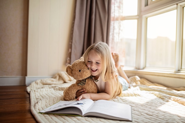 Foto nettes kleines kaukasisches mädchen in der freizeitkleidung, die ein buch mit ausgestopftem teddybärspielzeug liest