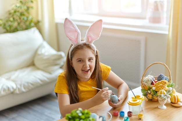 Nettes kleines Kind, das Hasenohren trägt. Mädchen sitzt sitzend am Tisch im Wohnzimmer. Kind malt Eier.