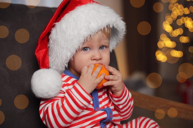 Nettes kleines Kind in Weihnachtsmütze mit reifer Mandarine im Sessel zu Hause