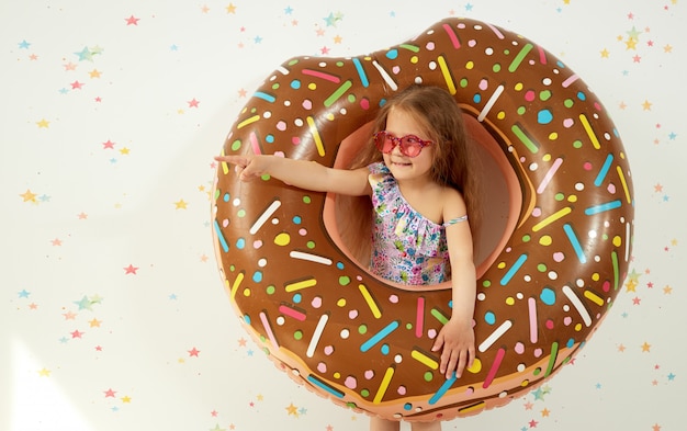 Nettes kleines Kindermädchen im Hut mit aufblasbarem Ring auf Farbwand. Quarantäne Sommerferien zu Hause