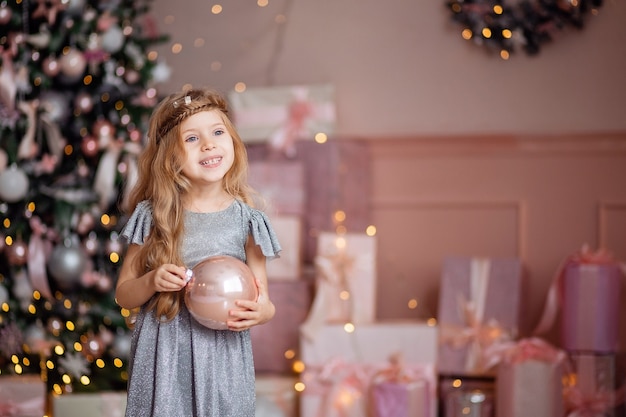 Nettes kleines lachendes blondes Mädchen mit langen Haaren spielt mit einem Weihnachtsball auf einem Hintergrund