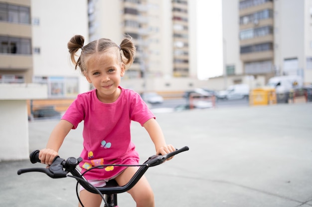 Nettes kleines lächelndes Mädchen, das Fahrrad in der Stadt fährt, am sonnigen Sommertag auf dem Parkplatz Aktive Familienfreizeit mit Kindern