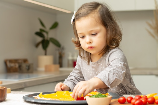Nettes kleines Mädchen 2-4 im grauen Kleid, das Pizza in der Küche kocht. Kid arrangiert Zutaten auf Pizzabasis
