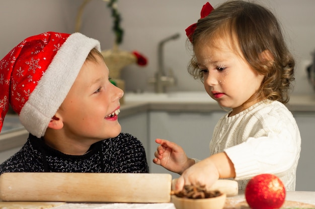 Nettes kleines Mädchen 2-4 mit roter Schleife und Junge 7-10 in einer Weihnachtsmütze, die Weihnachtslebkuchenplätzchen in der Küche des neuen Jahres macht.