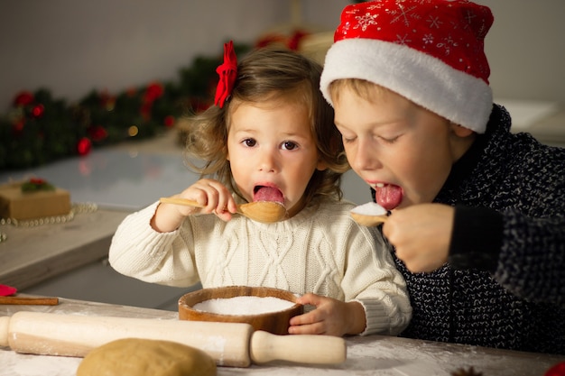 Nettes kleines Mädchen 2-4 mit roter Schleife und Junge 7-10 in einer Weihnachtsmütze, die Weihnachtslebkuchenplätzchen in der Küche des neuen Jahres macht.