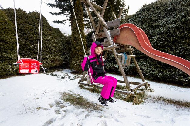 Foto nettes kleines mädchen, das am kalten verschneiten tag auf dem spielplatz schwingt