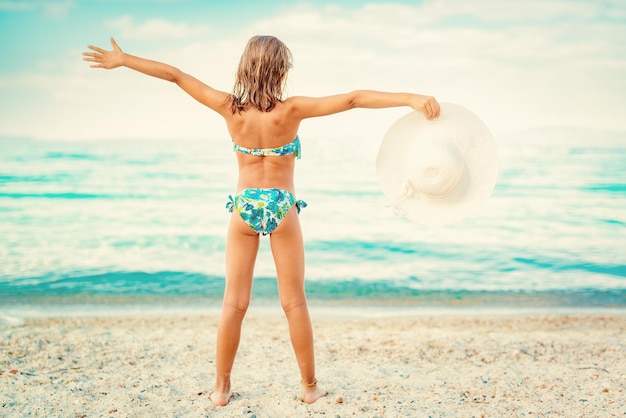 Nettes kleines Mädchen, das am Strand genießt. Sie poosiert und hält weißen Hut. Rückansicht.