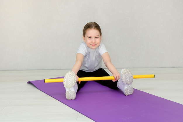 Nettes kleines Mädchen, das auf einer Matte sitzt und Übungen mit einem Gymnastikstock macht, scherzt Fitness