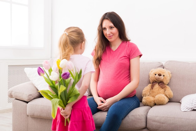 Nettes kleines Mädchen, das Blumen vor ihrer schwangeren Mutter hinter ihrem Rücken versteckt. Muttertagsfeier, Tochter, die ihrer Mutter gratuliert, Kopienraum.