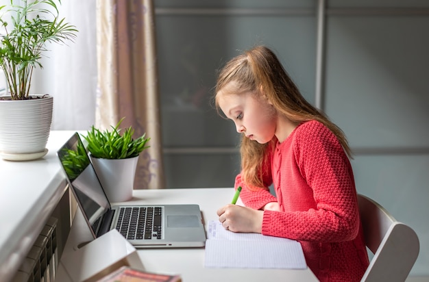 Nettes kleines Mädchen, das ernsthaft Hausaufgaben mit Laptop zu Hause für Online-Bildung, Heimstudium und Online-Kommunikation macht. Fernunterricht für Kinder. Bleib zuhause.