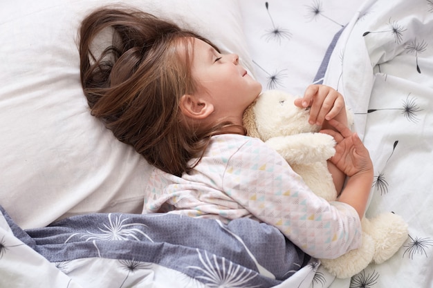 Nettes kleines Mädchen, das im Bett liegt, nach dem ganzen Tag spielend einschlafen, auf weißem Kissen unter Decke mit geschlossenen Augen liegend, charmantes Kind trägt Pyjama